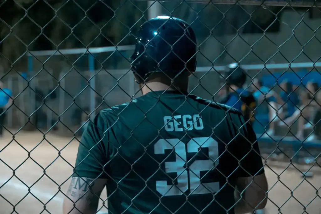 A baseball player wearing a helmet waits behind a fence during a night game