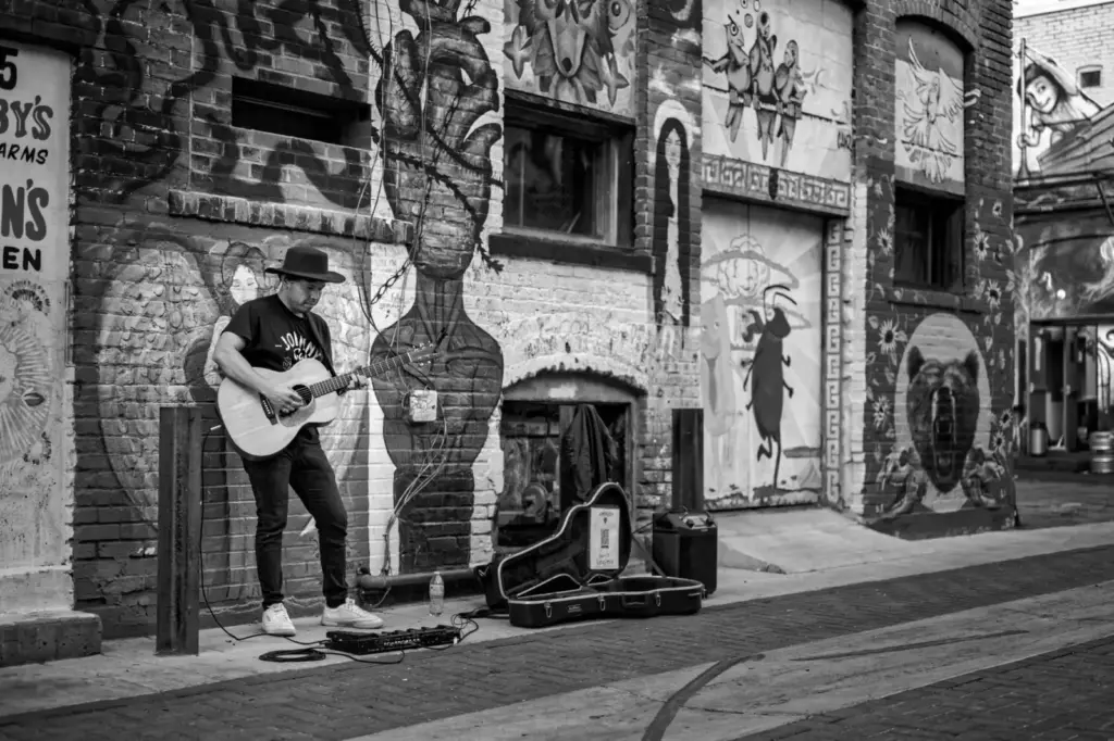 A black and white photo of a street musician playing guitar in front of mural-covered urban walls