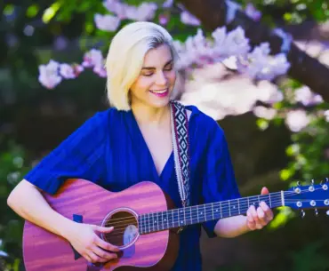 Blonde woman playing an acoustic guitar while sitting outdoors among blooming spring flowers