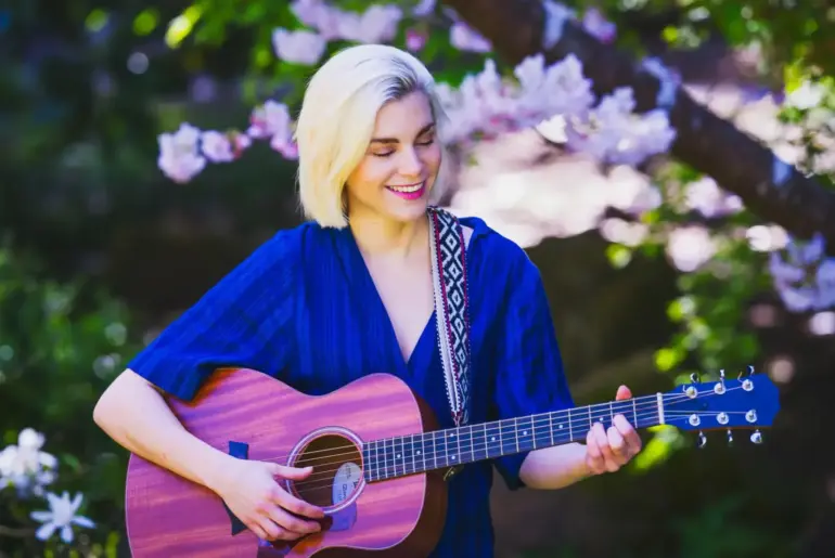 Blonde woman playing an acoustic guitar while sitting outdoors among blooming spring flowers