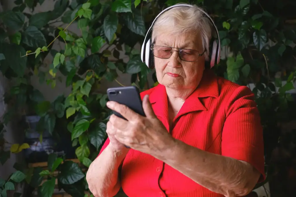 Concentrated senior female with headphones making video call online with mobile phone near fresh green leaves