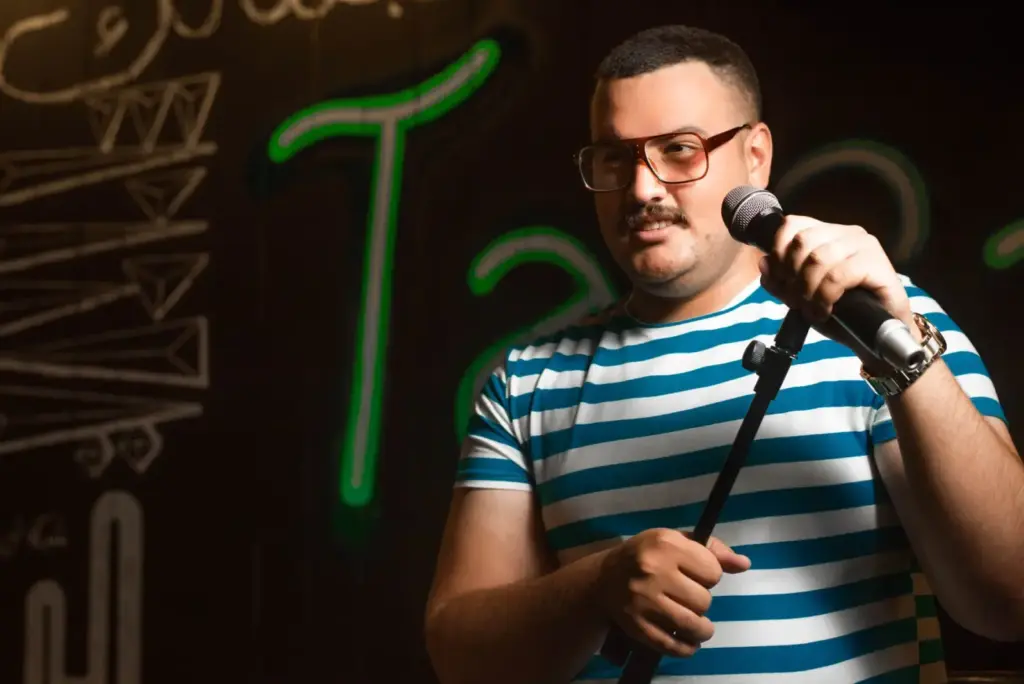 Man holding a microphone doing stand-up comedy in casual striped shirt on stage